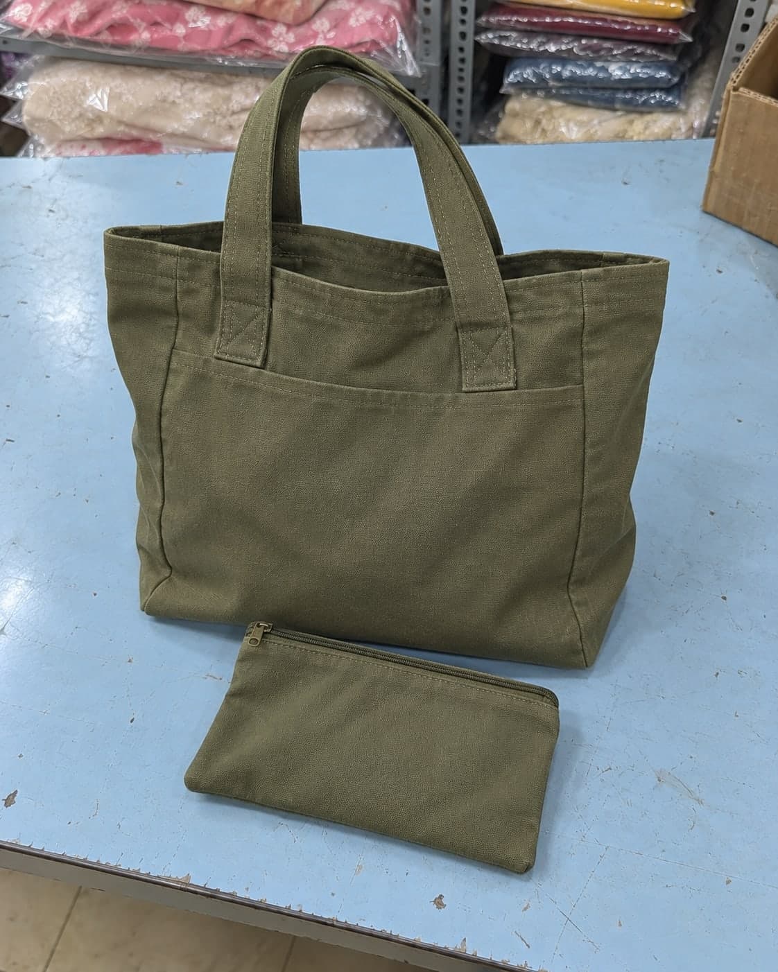An olive canvas tote bag and matching zip pouch on a shop counter