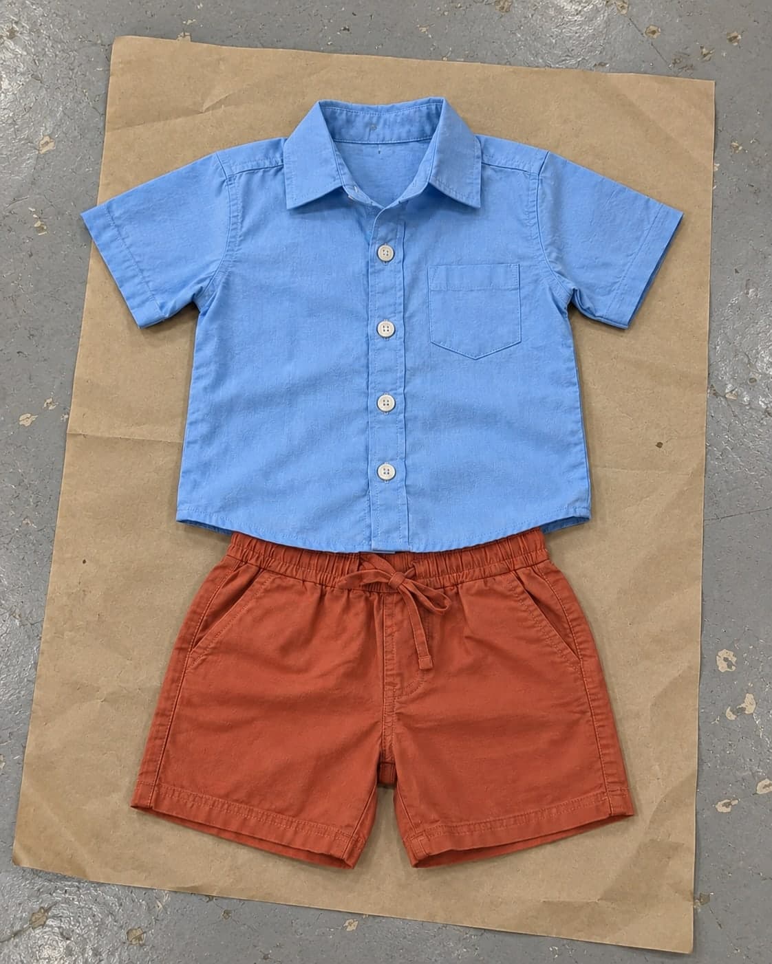 A child's blue cotton shirt and terracotta shorts laid out as an outfit