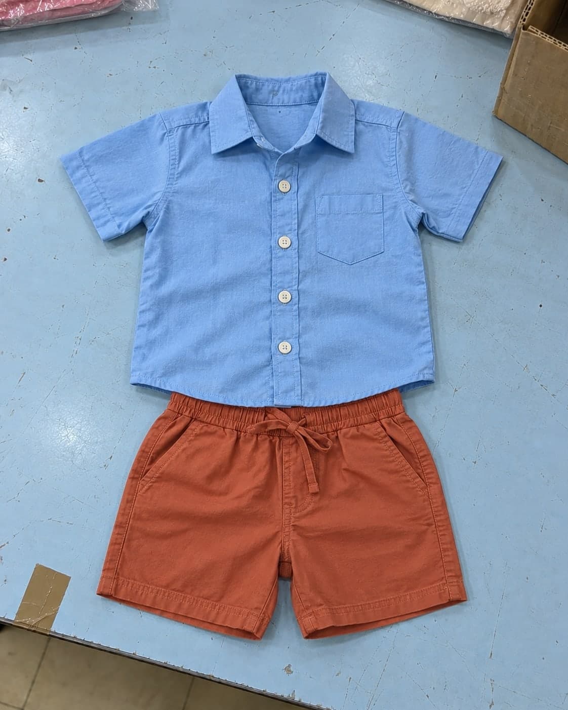A child's blue cotton shirt and terracotta shorts laid out as an outfit