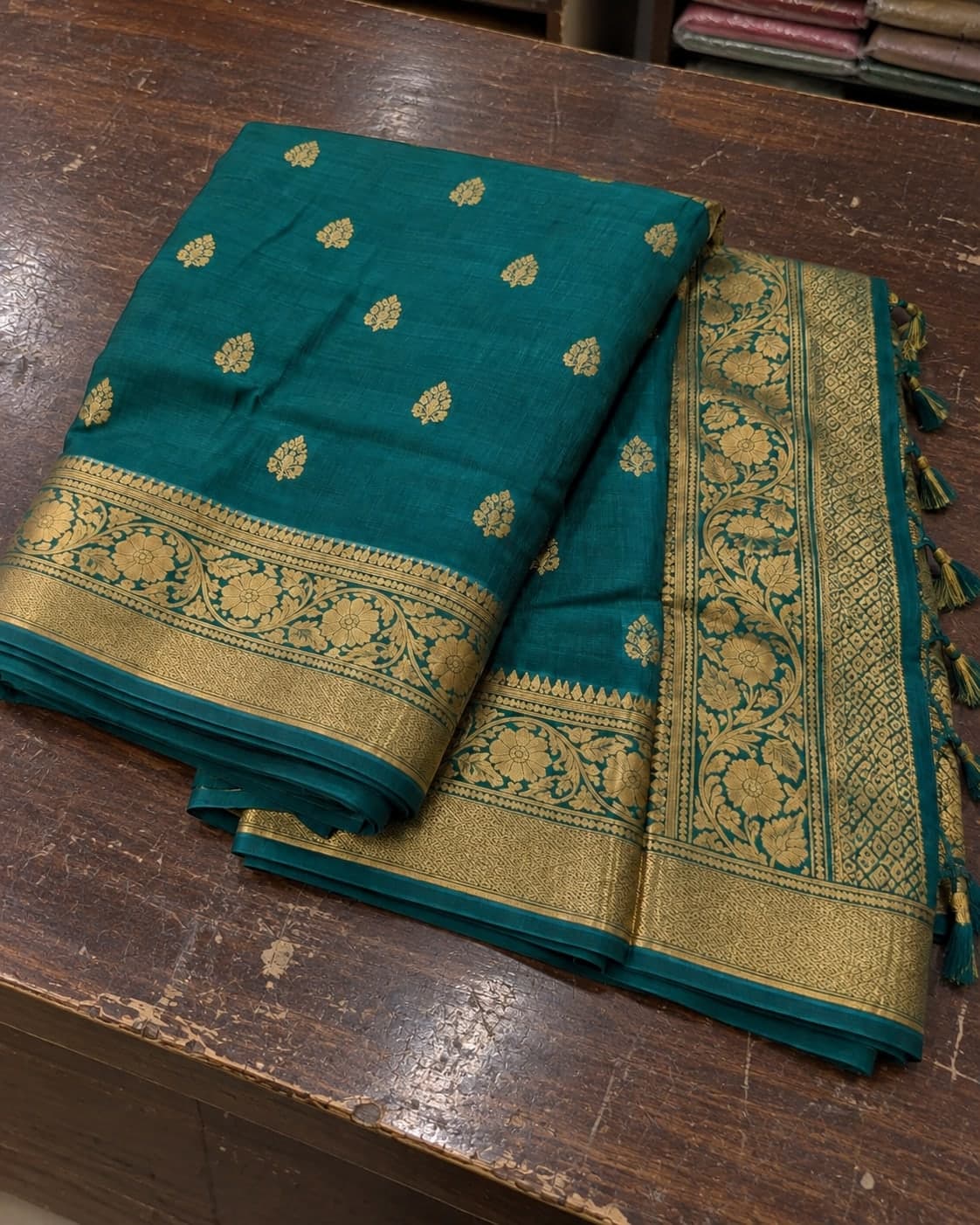A teal saree with a gold woven border folded on a shop counter