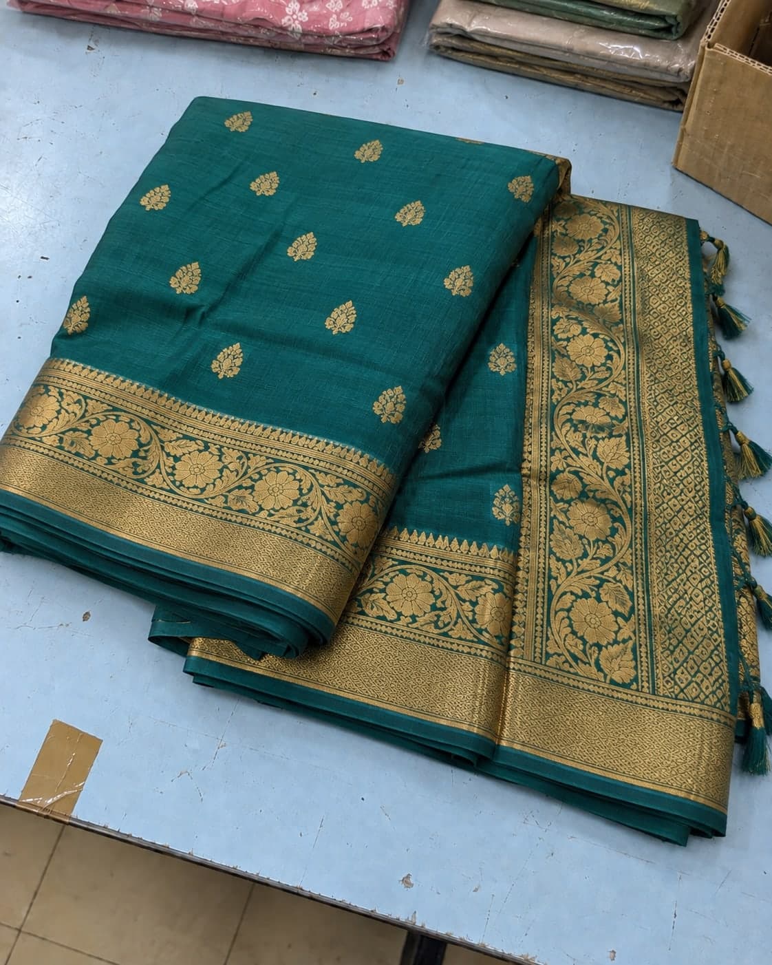 A teal saree with a gold woven border folded on a shop counter